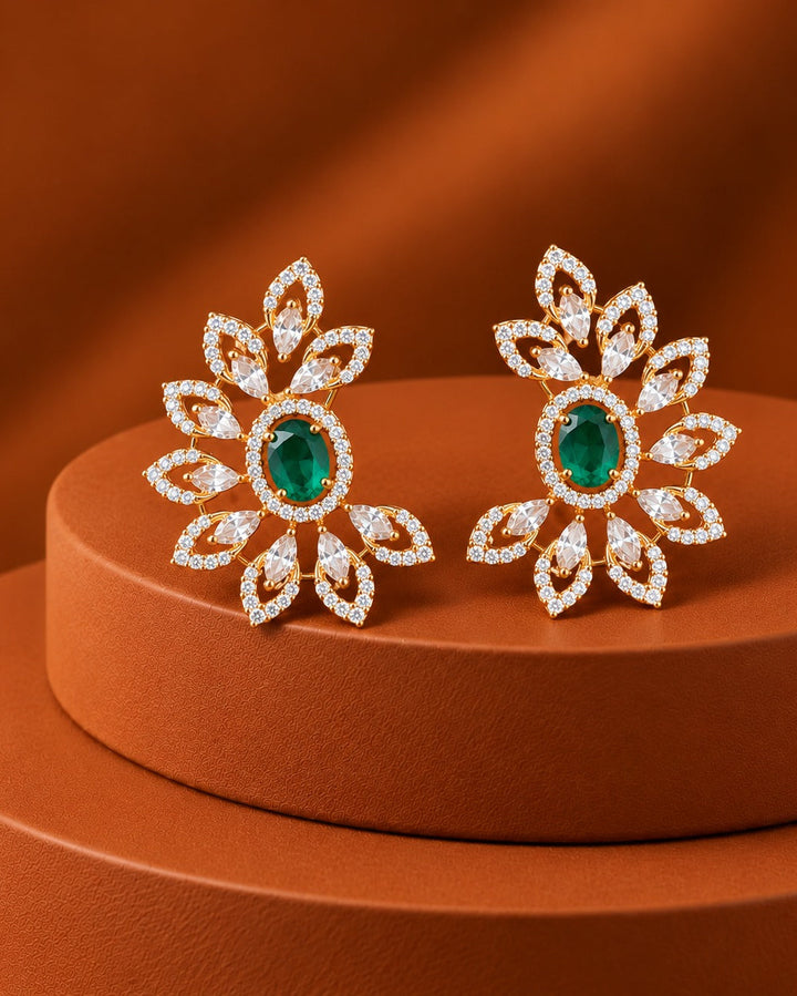 A pair of gold-toned floral stud earrings with oval green gemstones and pavé-set clear stones, displayed on a round terracotta pedestal against a matching warm brown background.