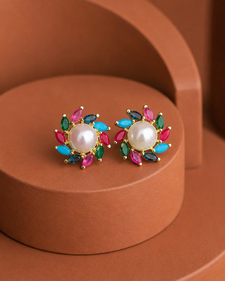 A pair of gold-toned floral stud earrings with a central white pearl and surrounding marquise-cut gemstones in pink, turquoise, and dark blue, displayed on a terracotta-colored circular pedestal.