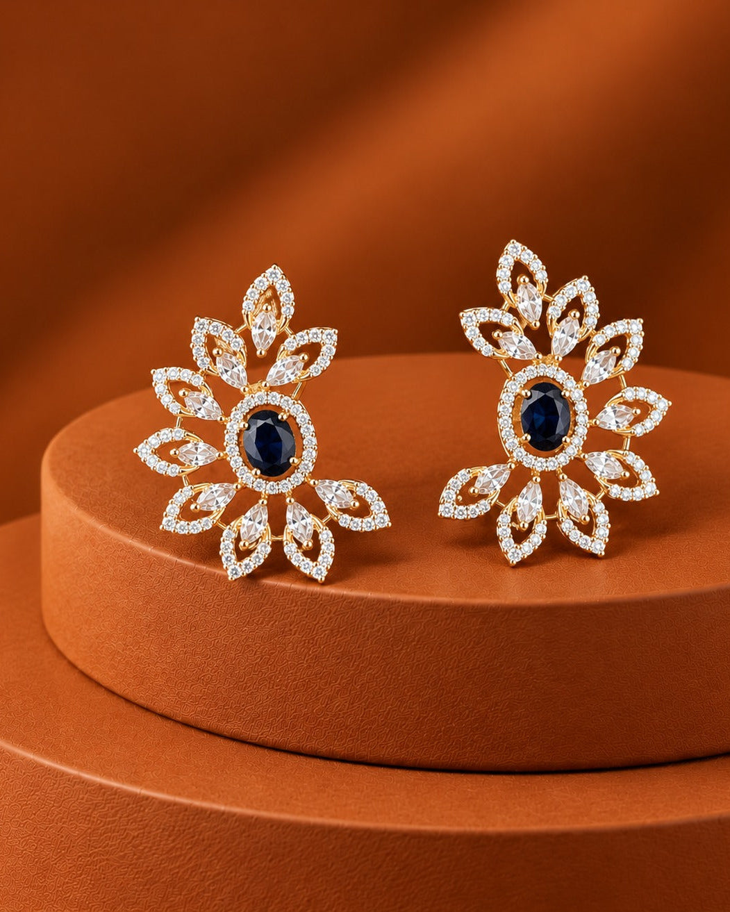 A pair of gold-toned floral stud earrings with oval dark blue center stones and surrounding clear stones, displayed on a round brown pedestal against a warm brown background.