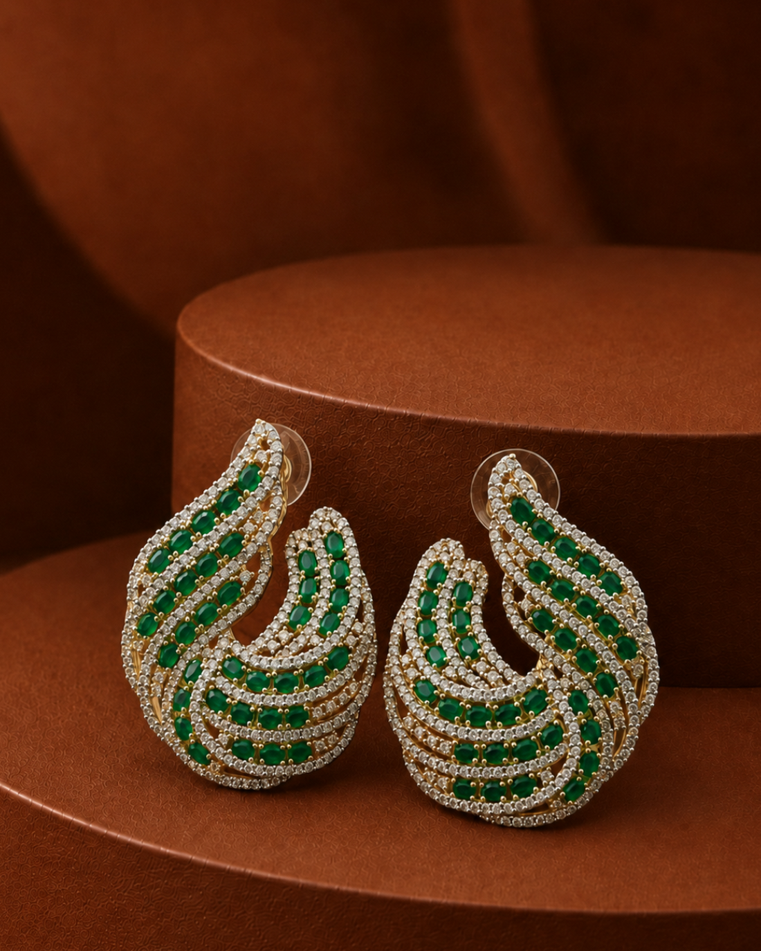 A pair of gold-toned huggie hoop earrings embellished with multiple rows of green emeralds and clear pavé stones, displayed on a brown textured pedestal.