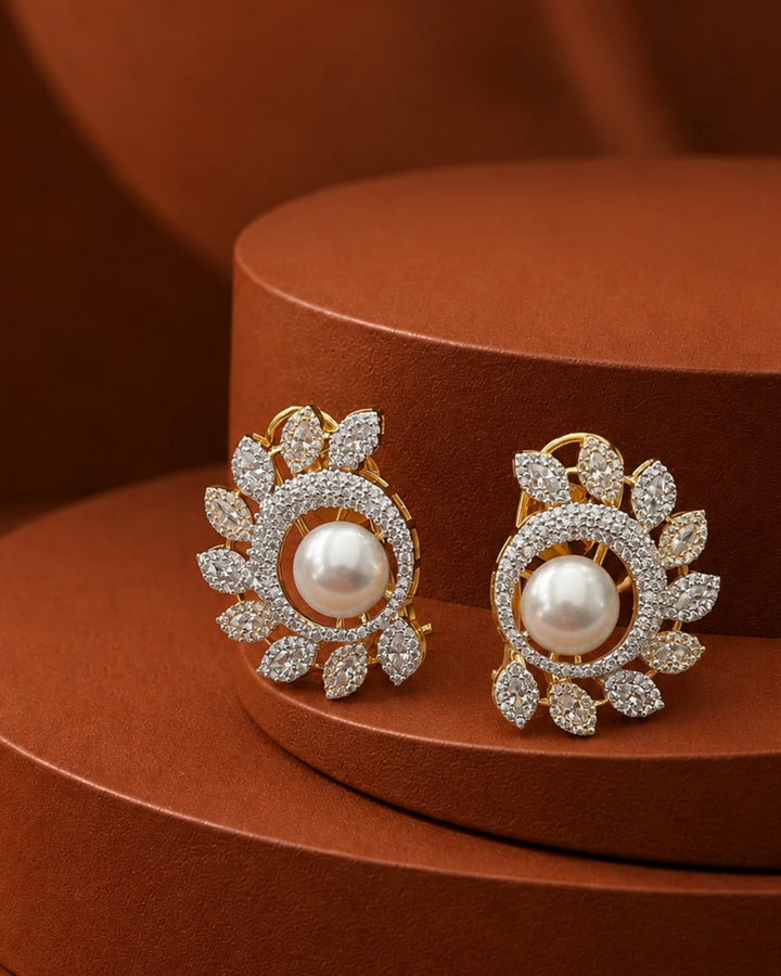 A pair of gold-toned leaf-shaped earrings with central white pearls and surrounding pavé-set clear stones, displayed on a round brown pedestal against a matching brown background.