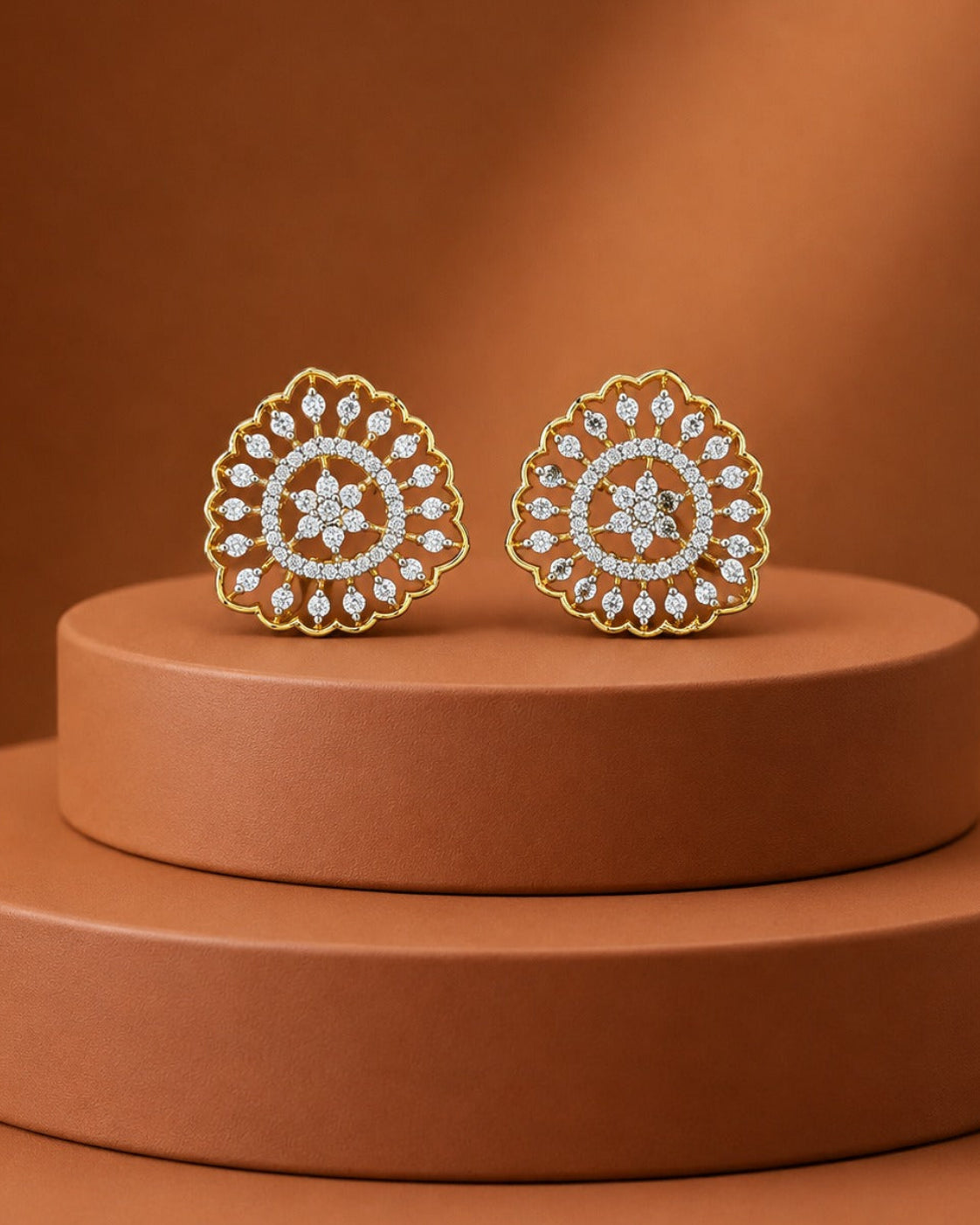 A pair of gold-toned mandala stud earrings with clear pavé stones, displayed on a tiered terracotta pedestal against a matching warm brown background.