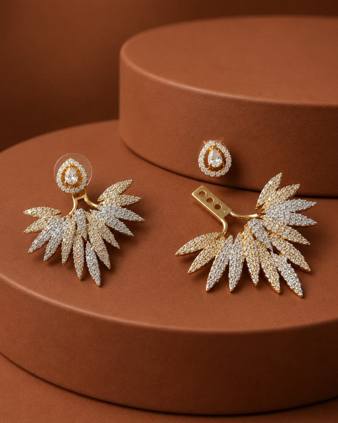 A pair of gold-toned statement earrings with leaf-shaped pavé crystal clusters and matching oval stud backings, displayed on a warm brown cylindrical pedestal.