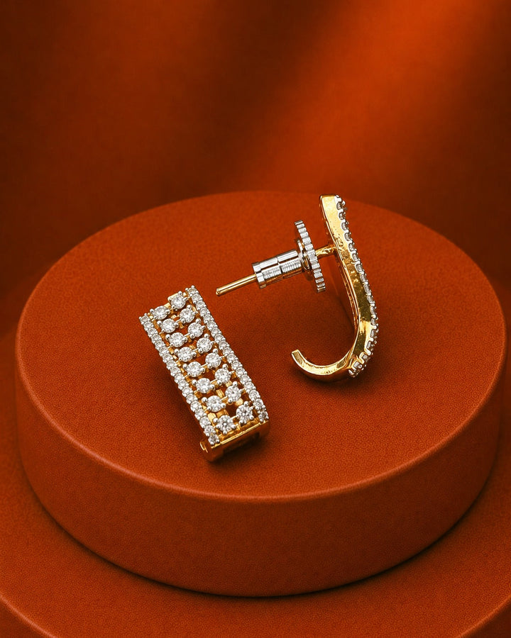 A pair of gold-toned statement earrings displayed on a round orange-brown pedestal. One earring is shown front-facing as a double-row rectangular bar set with clear stones, and the other is shown in profile as a curved hook with a row of clear stones along the front.