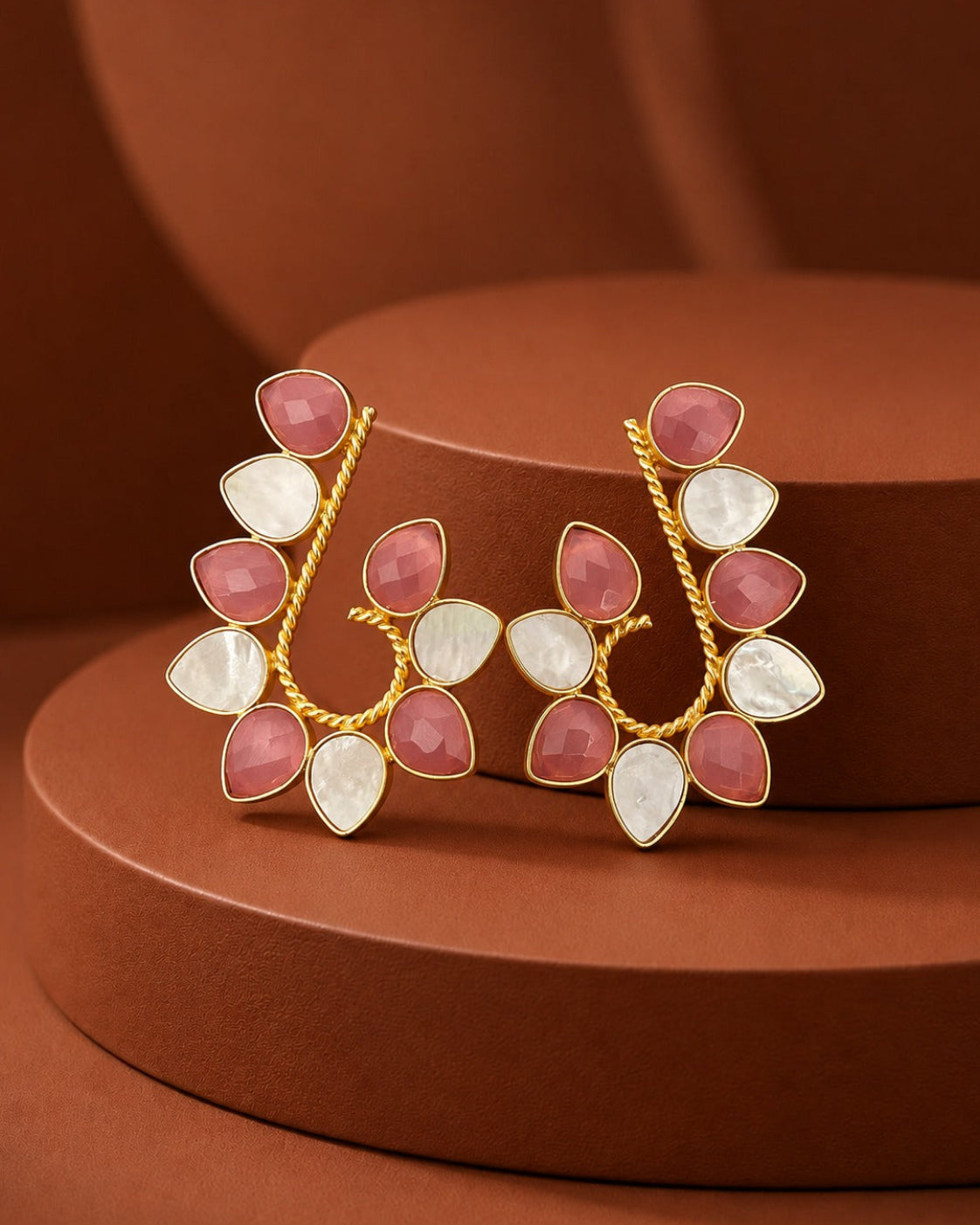 A pair of gold-toned statement earrings with alternating pink teardrop-shaped stones and white pear-shaped stones, displayed on a brown tiered pedestal.