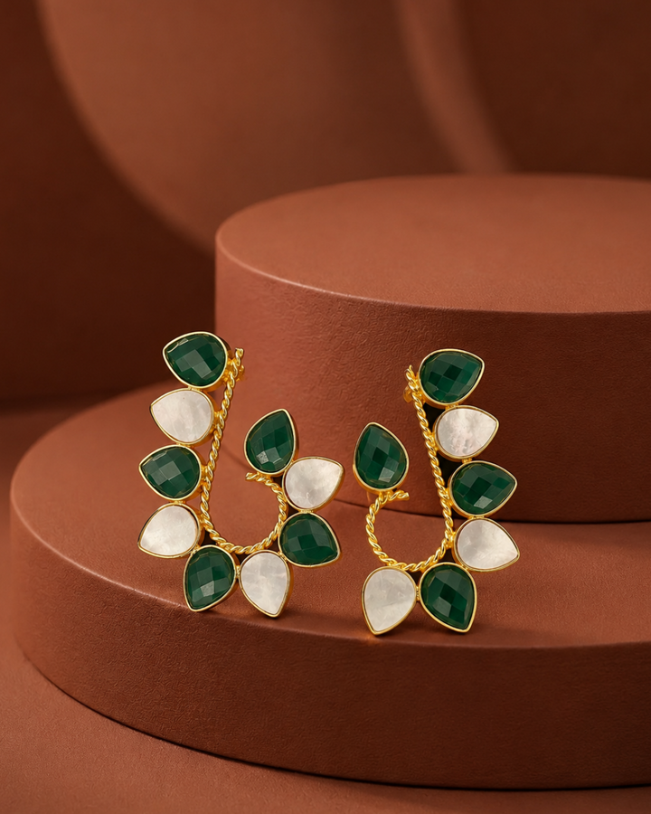 A pair of gold-toned statement earrings with faceted green and white teardrop-shaped stones, displayed on a brown cylindrical pedestal.