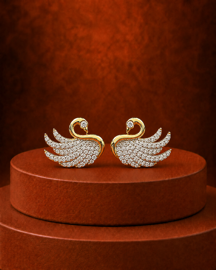 A pair of gold-toned swan-shaped earrings paved with clear stones, displayed on a round brown pedestal against a textured brown background.