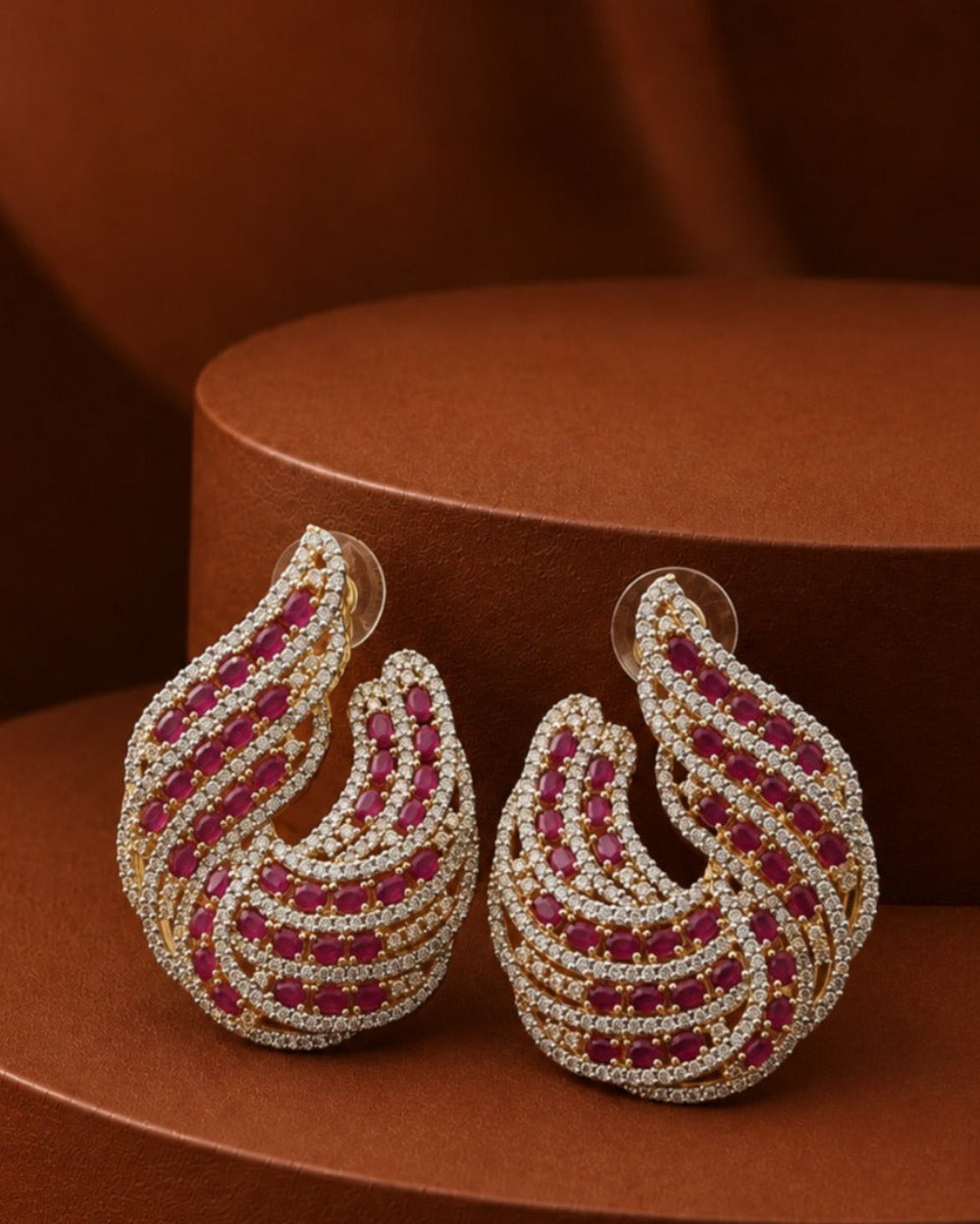A pair of gold-toned teardrop-shaped earrings paved with small clear stones and set with curved rows of deep red gemstones, displayed on a brown textured pedestal.