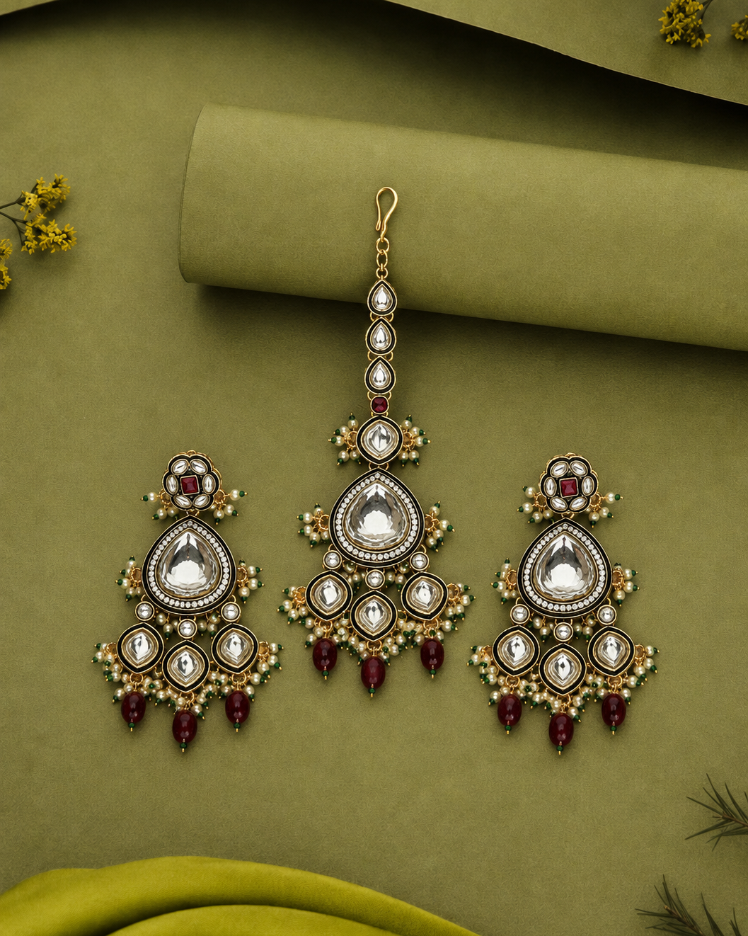 A pair of gold-toned traditional Indian choker earrings with matching drop earrings, decorated with white stones, small green beads, and deep red teardrop beads, displayed on an olive green textured background.
