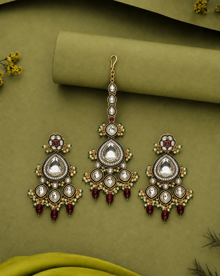 A pair of gold-toned traditional Indian choker earrings with matching drop earrings, decorated with white stones, small green beads, and deep red teardrop beads, displayed on an olive green textured background.
