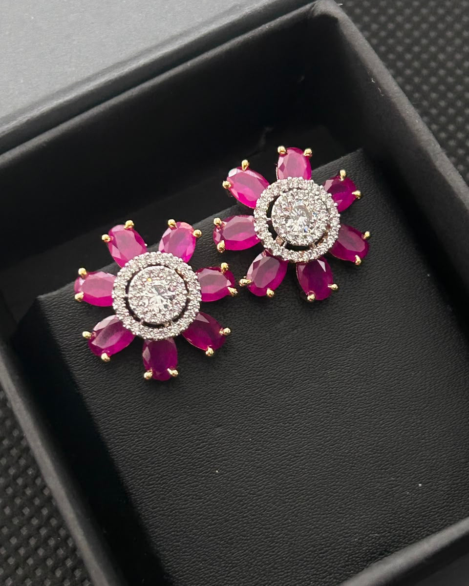 A pair of gold and crystal stud earrings with bright pink gemstone petals, displayed on a black textured jewelry box.
