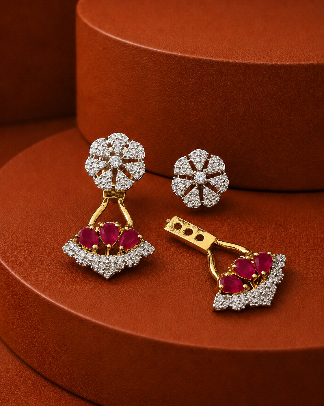 A pair of gold and diamond drop earrings with red gemstone accents, displayed on a textured brown surface with a matching brown cylindrical prop.