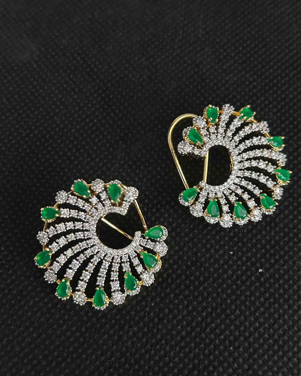 A pair of gold and diamond drop earrings with green emerald-cut accents, displayed on a textured black background.
