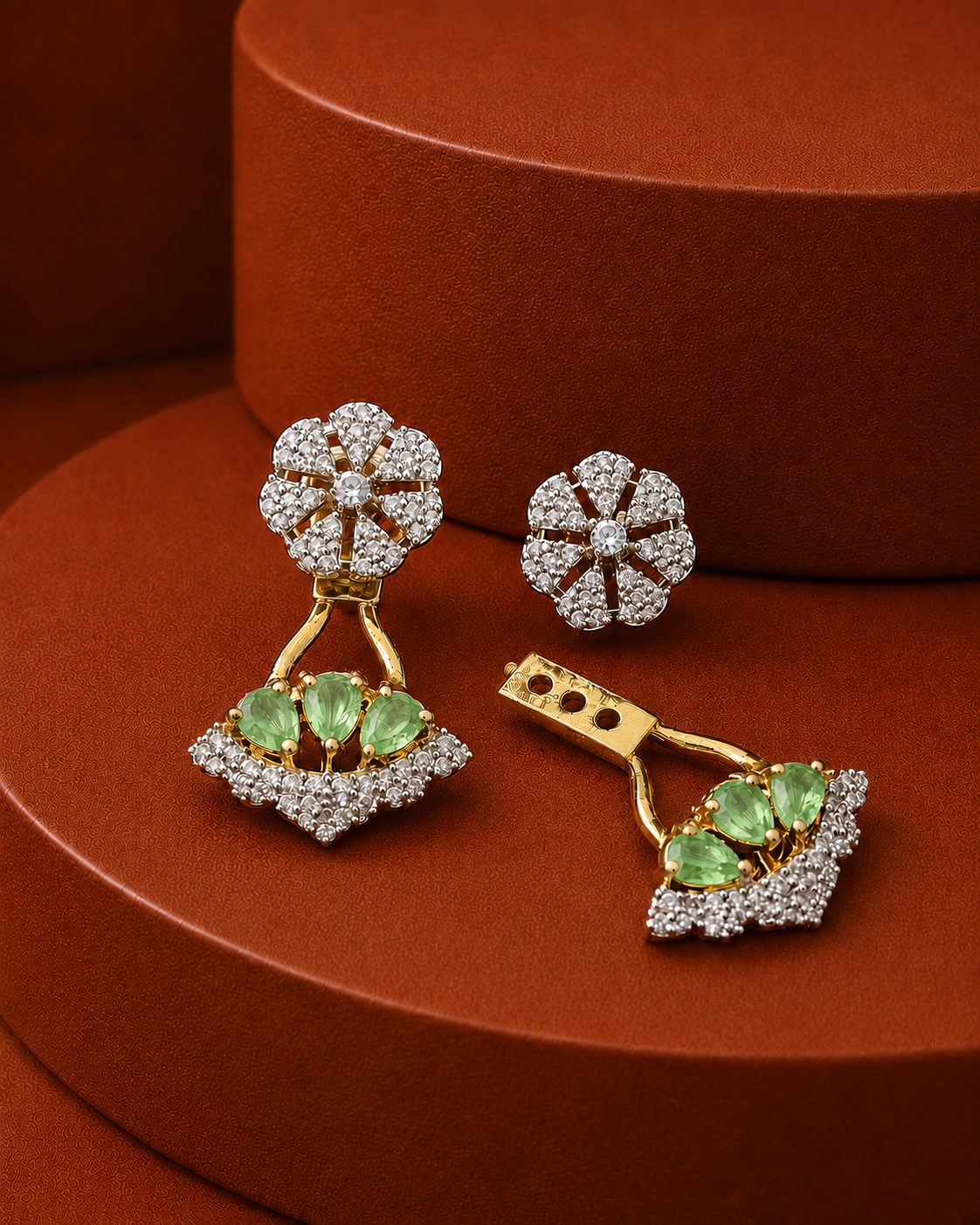 A pair of gold and diamond drop earrings with green gemstone accents, displayed on a textured brown circular pedestal.