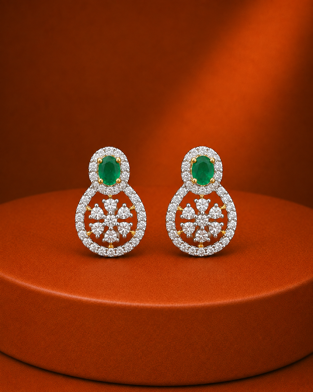 A pair of gold and diamond drop earrings with oval green emerald-cut center stones, displayed on a round orange-brown pedestal against a matching orange-brown background.