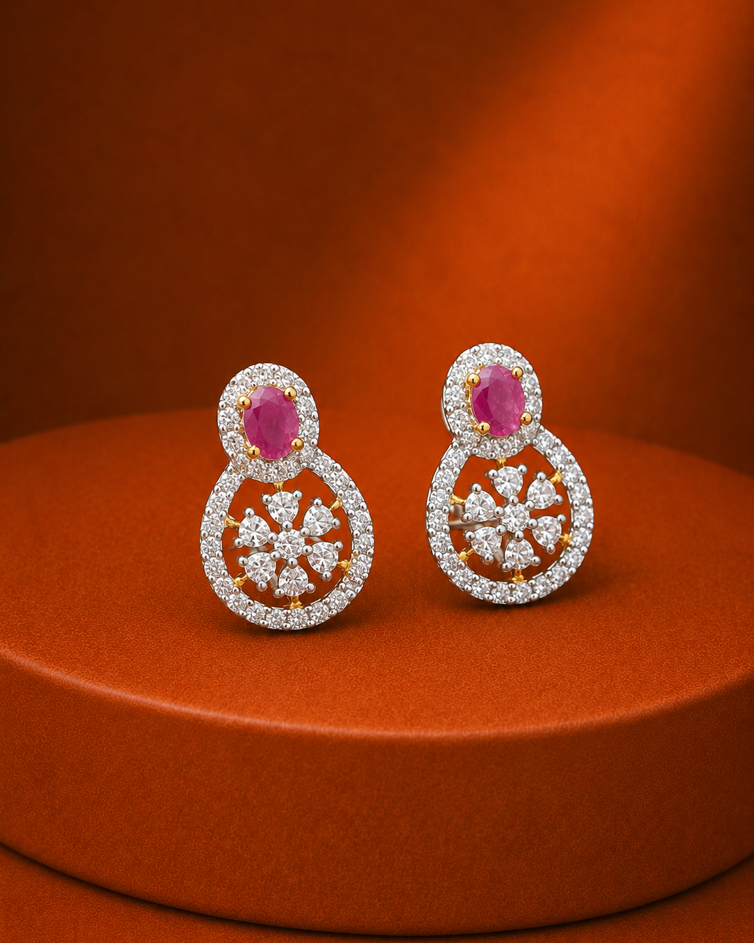 A pair of gold and diamond drop earrings with oval pink gemstones, displayed on a round orange pedestal against a matching orange background.