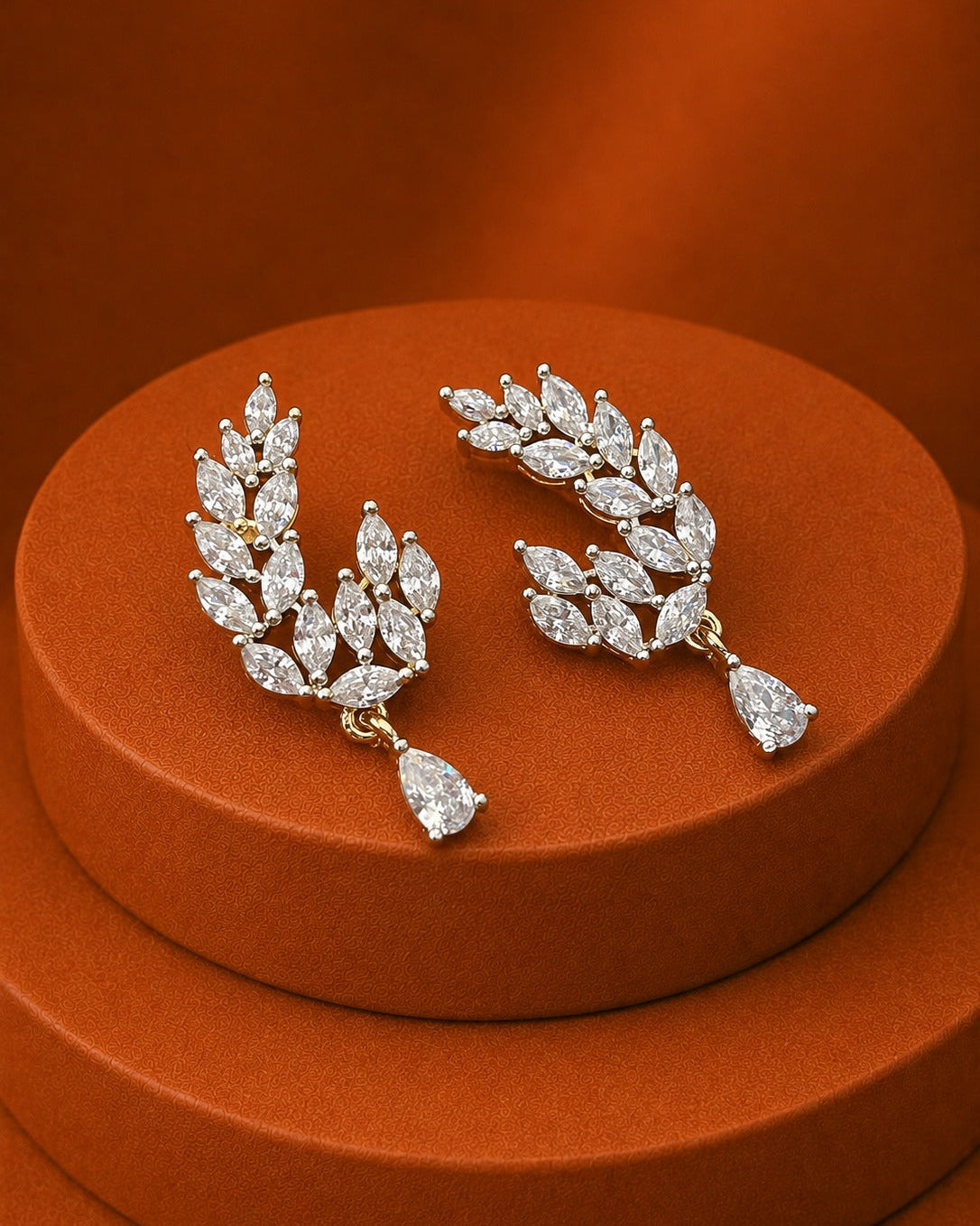 A pair of gold and diamond leaf-shaped drop earrings displayed on a round terracotta pedestal against a matching terracotta background.