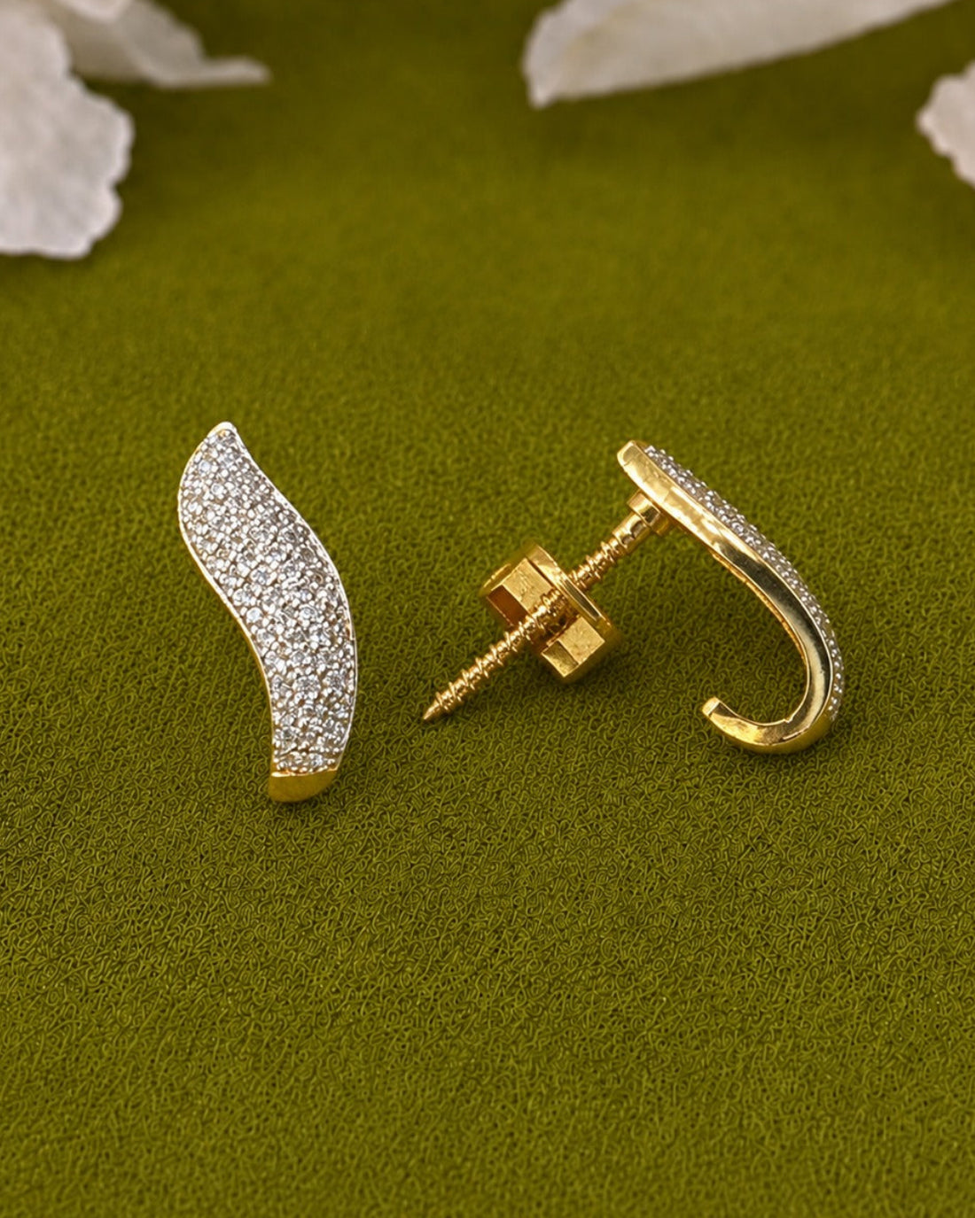 A pair of gold and diamond pavé earrings displayed on a textured olive green surface with white flower petals in the background. One earring is shown from the front, displaying the pavé diamond setting, and the other is turned to show the post and screw-back clasp.