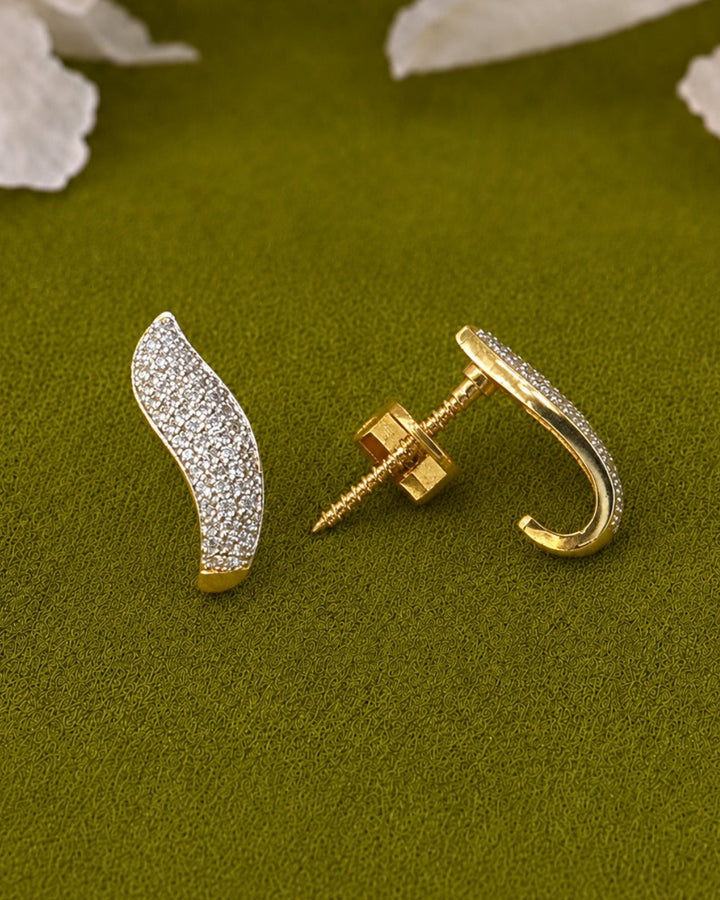 A pair of gold and diamond pavé earrings displayed on a textured olive green surface with white flower petals in the background. One earring is shown from the front, displaying the pavé diamond setting, and the other is turned to show the post and screw-back clasp.