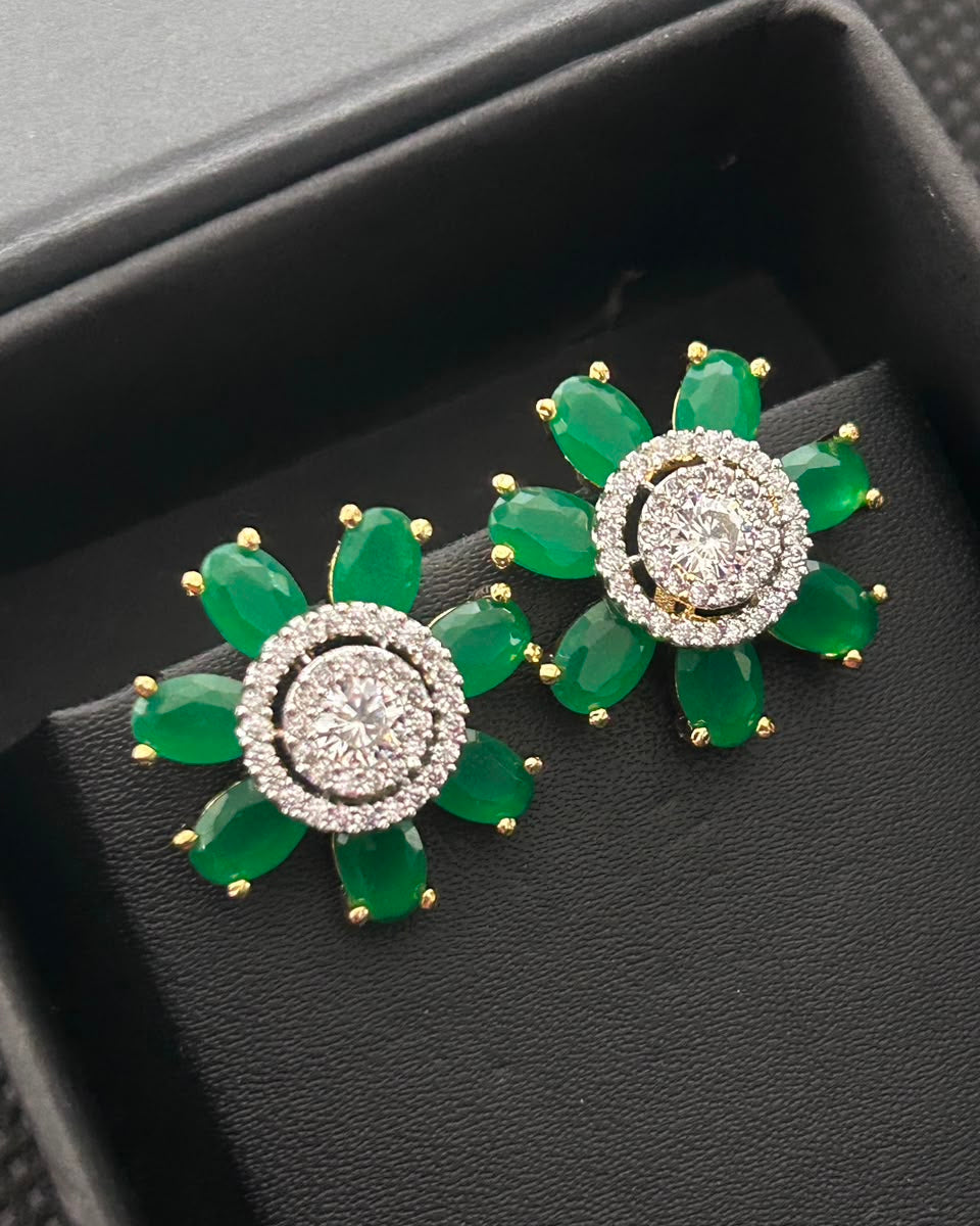 A pair of gold and green gemstone flower-shaped earrings displayed on a black jewelry box. Each earring features a central round cluster of clear stones surrounded by green marquise-cut stones, with gold prongs holding the outer petals.