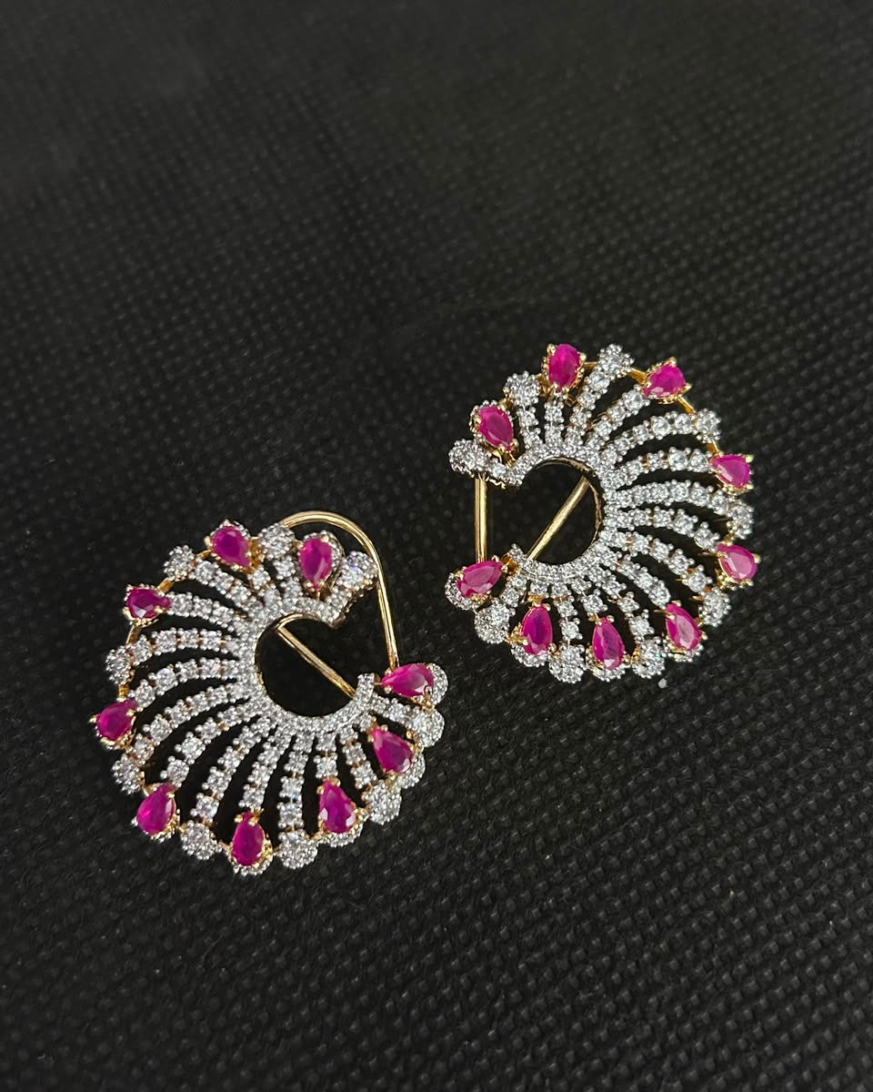 A pair of gold and silver-tone hoop earrings decorated with clear pavé stones and bright pink teardrop gemstones, displayed on a textured black background.