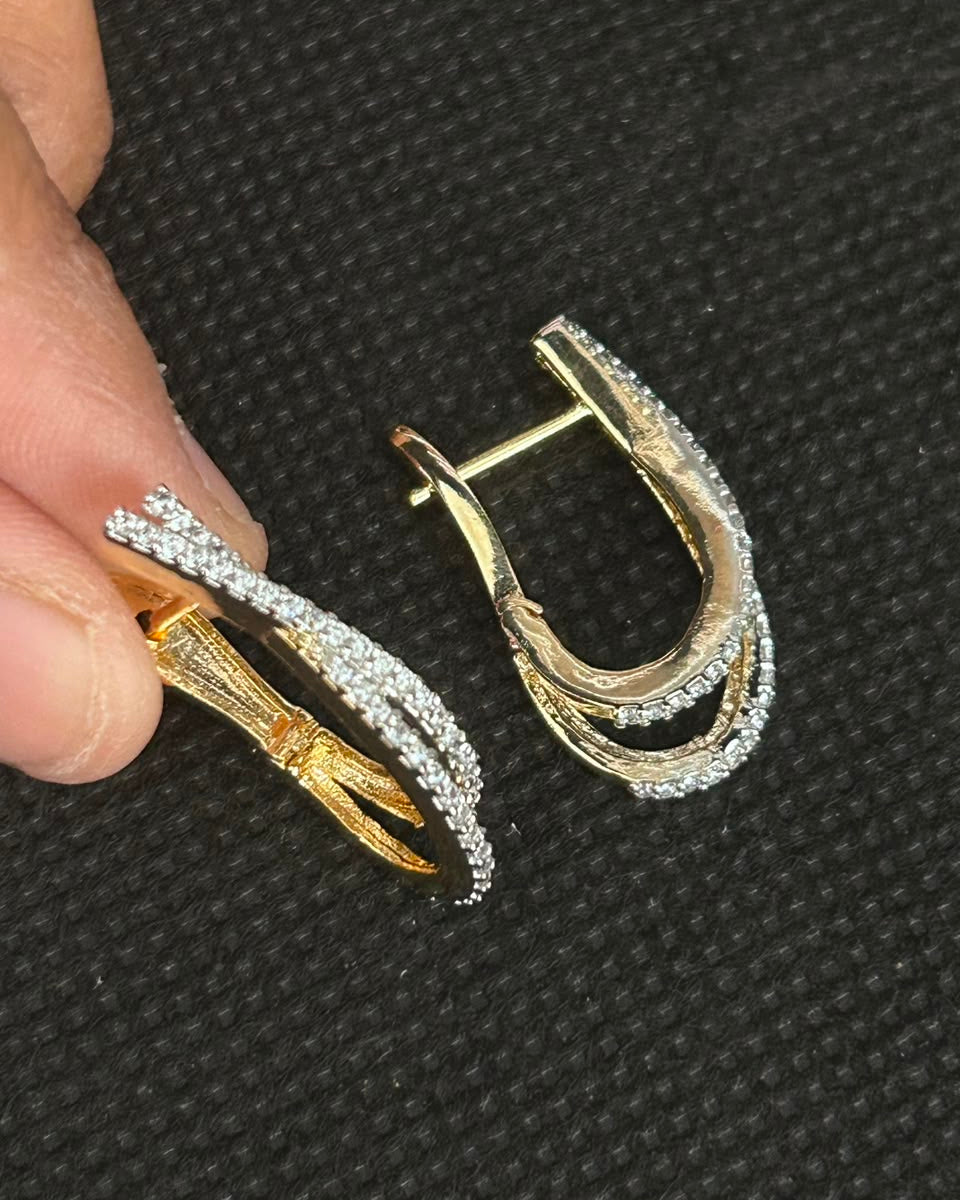 A pair of gold and silver hoop earrings with pavé-set clear stones, shown with one earring held by fingers against a black textured background.