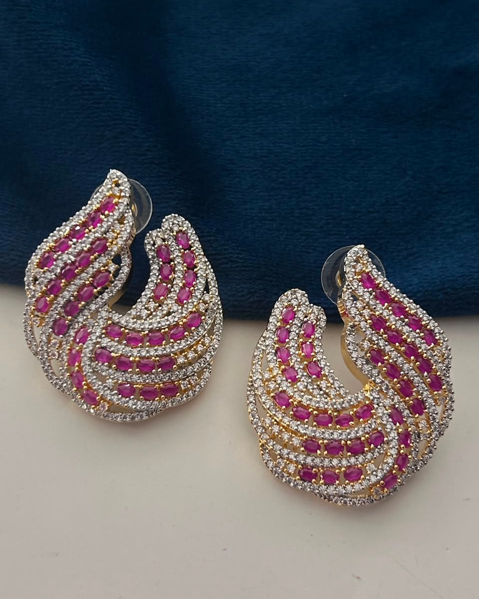 A pair of gold and silver  hoop earrings adorned with rows of pink gemstones and clear rhinestones, displayed on a light surface with a dark blue fabric background.