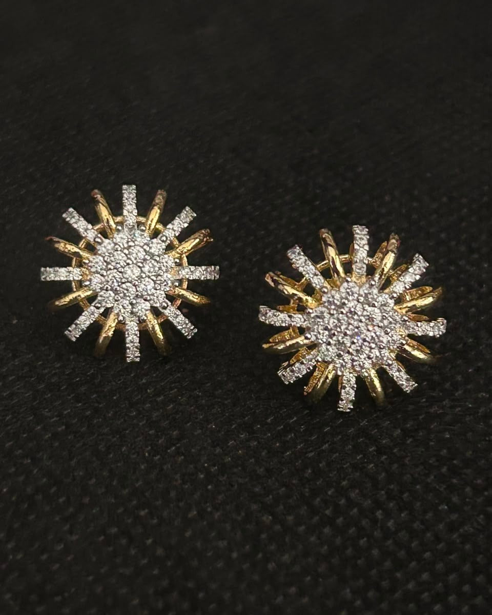 A pair of gold and silver starburst stud earrings paved with clear stones, displayed on a textured black background.