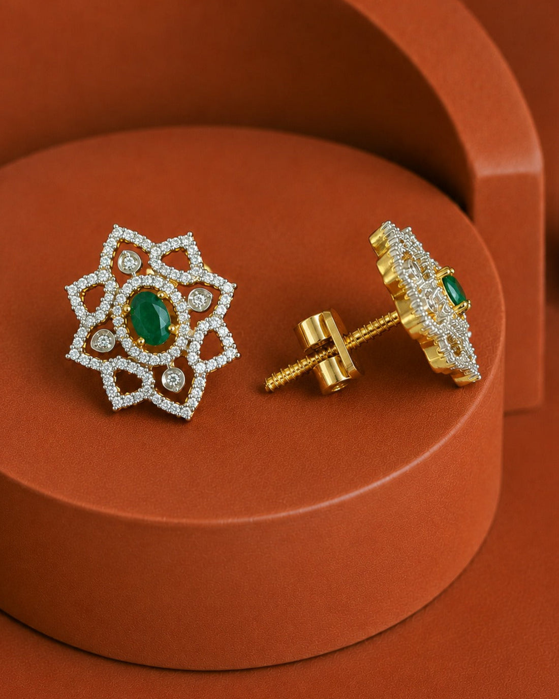 A pair of gold and white gold post earrings with green emerald-cut center stones and pavé-set clear stones, displayed on a terracotta-colored geometric pedestal.