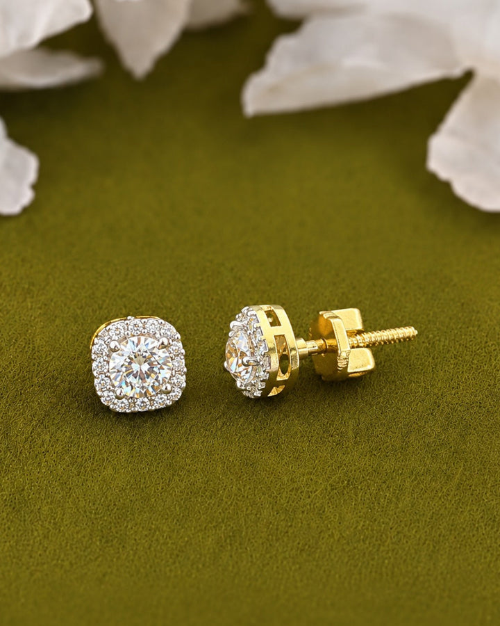 A pair of gold cushion-cut stud earrings with clear pavé-set stones, shown on an olive green textured background with soft white flower petals in the background.