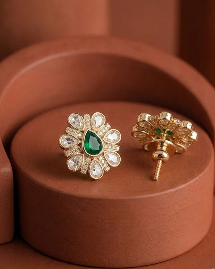 A pair of gold floral stud earrings with a central green teardrop gemstone and surrounding clear stones, displayed on a terracotta-colored circular pedestal.