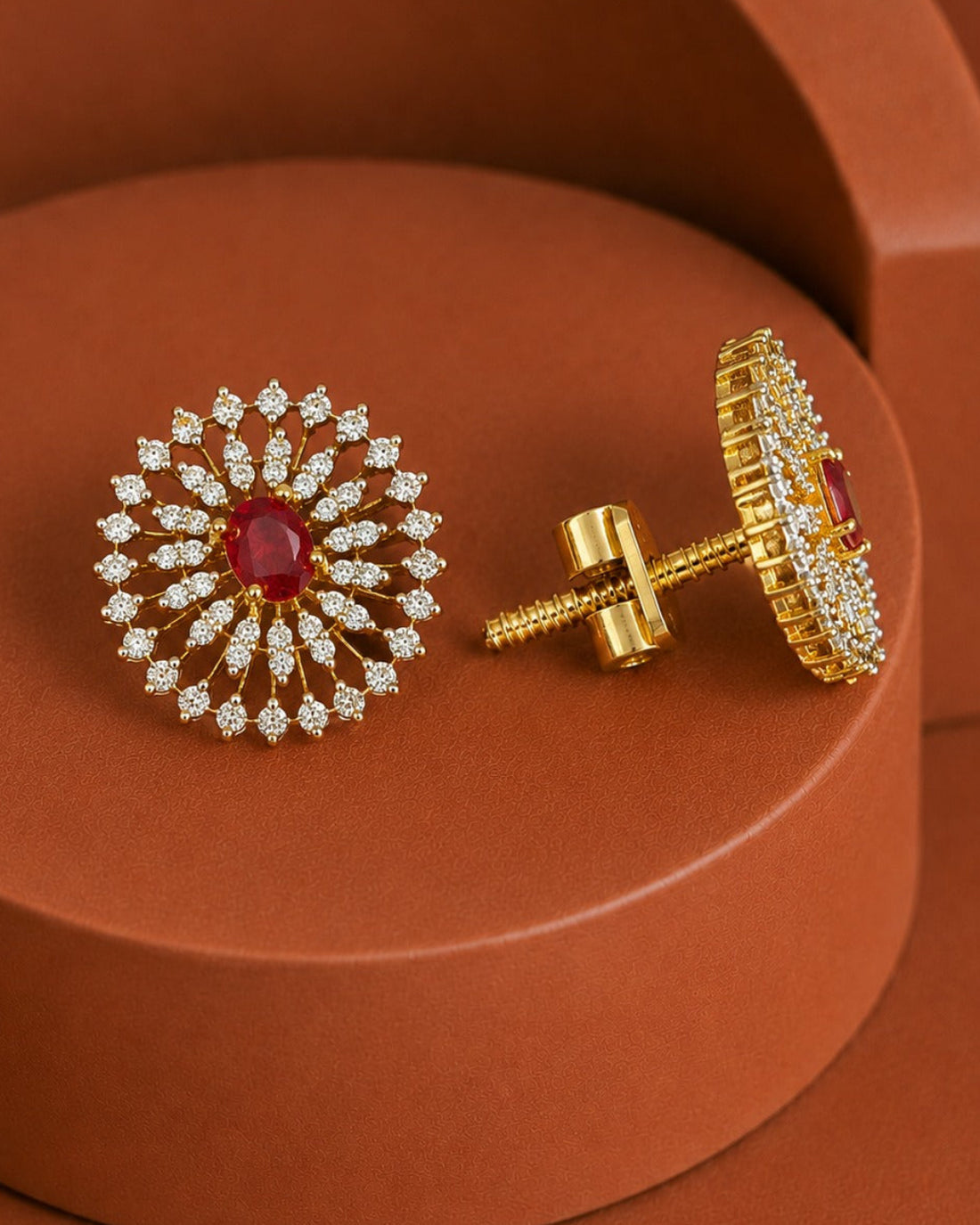 A pair of gold floral stud earrings with a central red gemstone and surrounding clear stones, displayed on a terracotta-colored geometric pedestal.