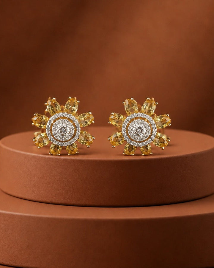 A pair of gold flower-shaped stud earrings with yellow gemstones and a central halo of clear stones, displayed on a round brown pedestal against a warm brown background.