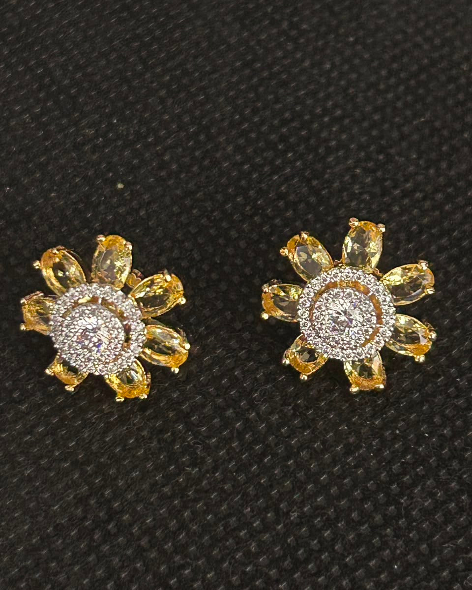 A pair of gold flower-shaped stud earrings with yellow marquise-cut petals and a clear pavé center, displayed on a textured black background.