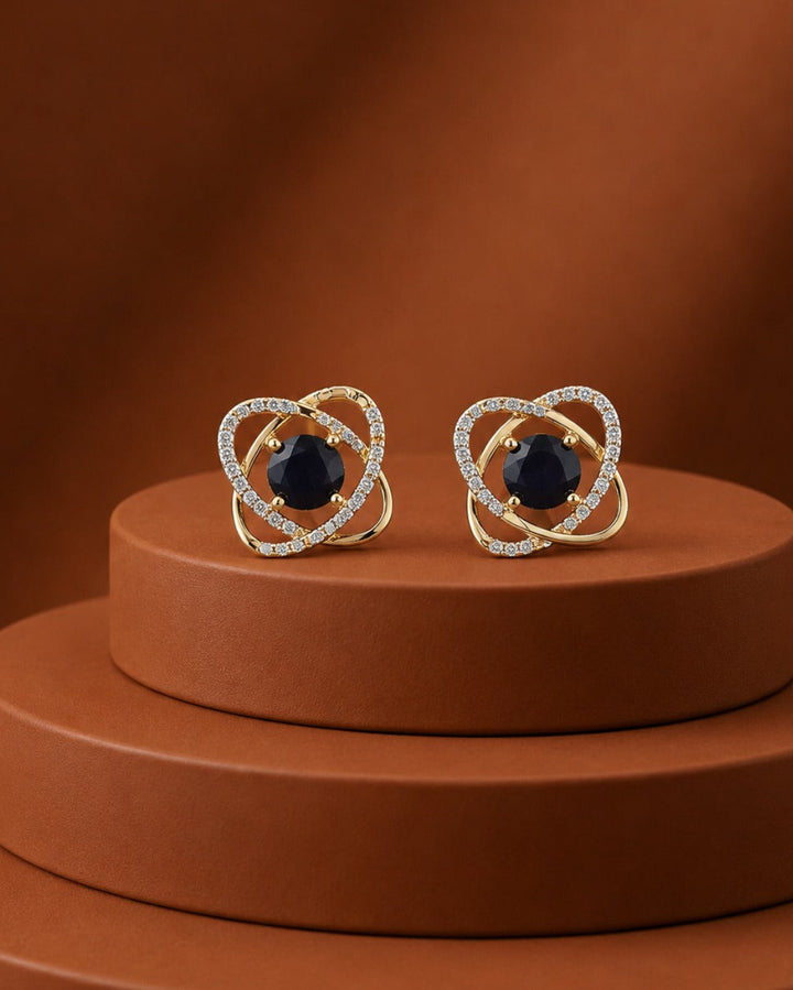 A pair of gold heart-shaped stud earrings with central dark blue gemstones and rows of small clear stones, displayed on a brown tiered pedestal against a warm brown background.
