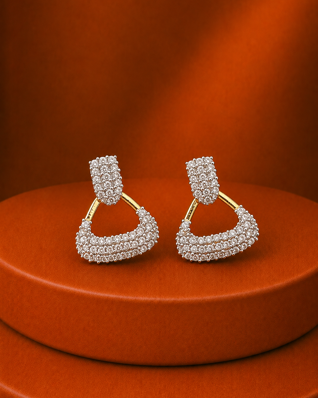 A pair of gold hoop earrings with pavé-set clear stones, displayed on a round orange pedestal against a matching orange background.