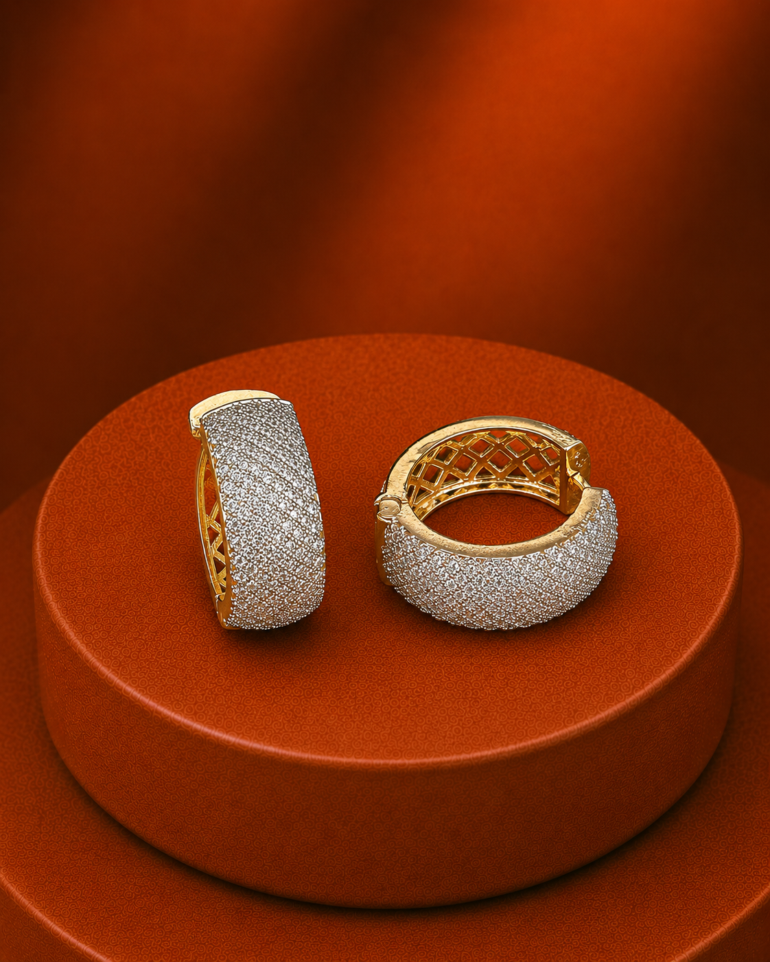 A pair of gold huggie hoop earrings covered in pavé-set clear stones, displayed on a round terracotta pedestal against a matching warm brown background.