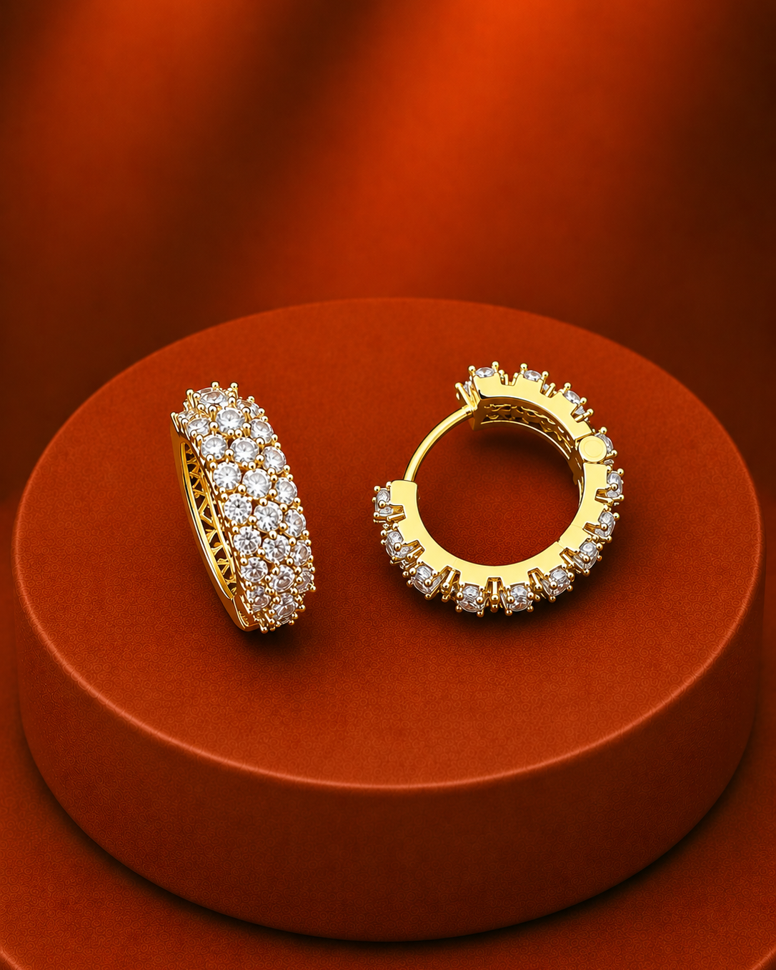 A pair of gold huggie hoop earrings paved with clear cubic zirconia stones, displayed on a round orange-brown pedestal against a matching orange-brown background.