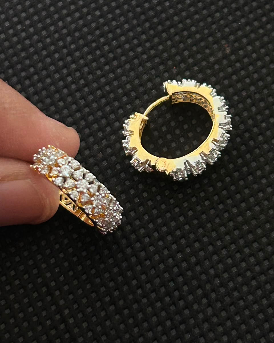 A pair of gold huggie hoop earrings paved with clear cubic zirconia stones, shown with one earring on a textured black background and the other held between fingers to demonstrate scale.