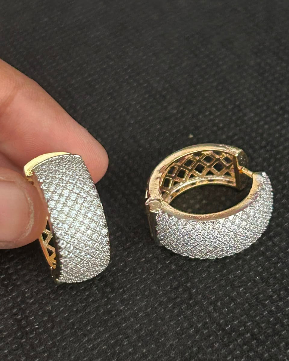 A pair of gold huggie hoop earrings paved with clear stones, held by a hand against a dark textured background.