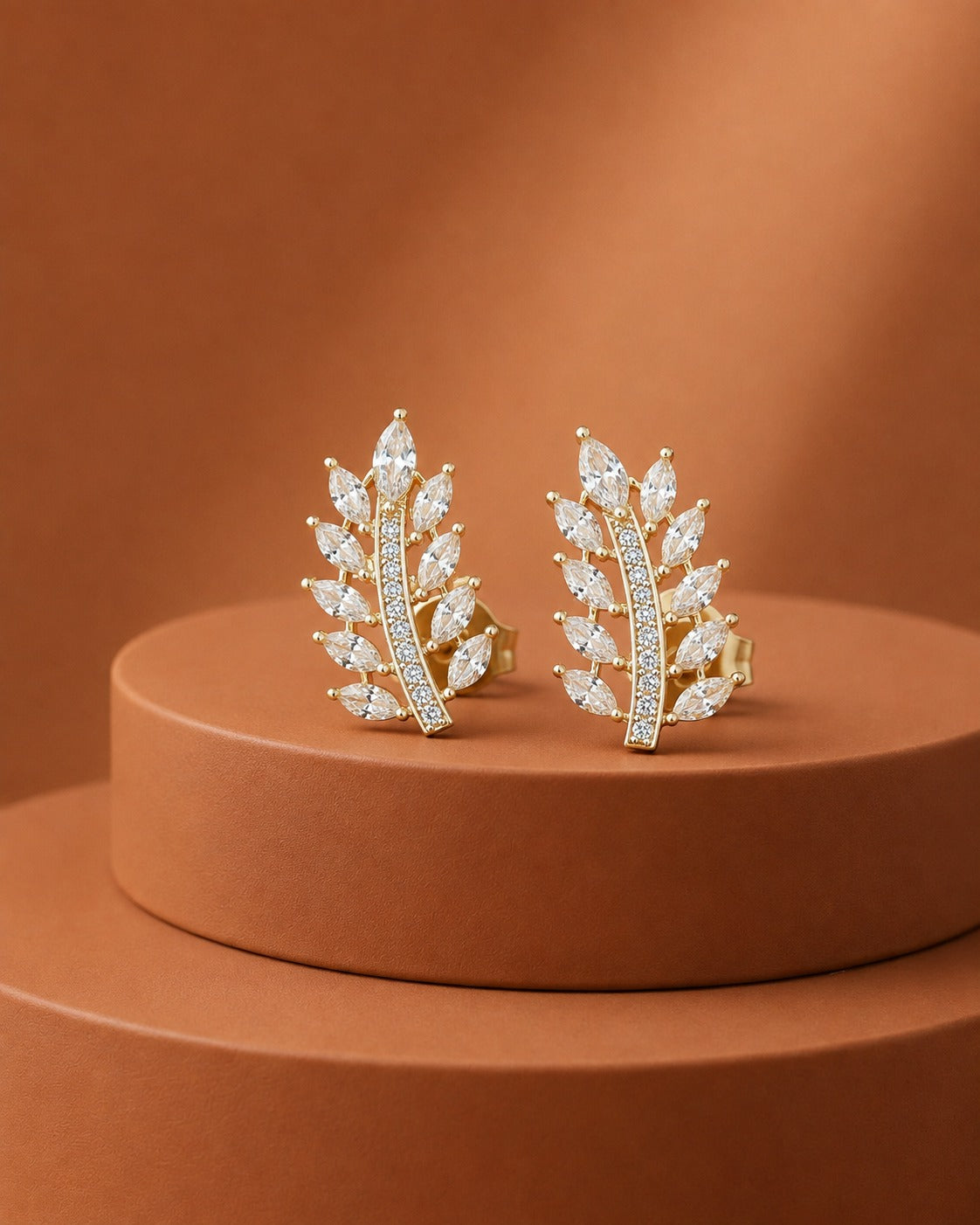A pair of gold leaf-shaped stud earrings set with marquise-cut clear stones, displayed on a round terracotta pedestal against a matching warm brown background.