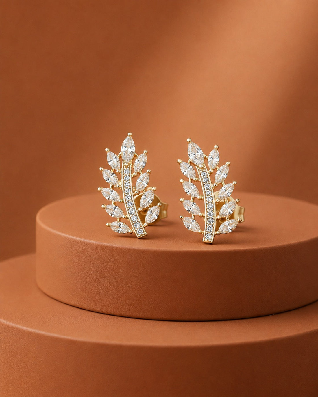 A pair of gold leaf-shaped stud earrings set with marquise-cut clear stones, displayed on a round terracotta pedestal against a matching warm brown background.