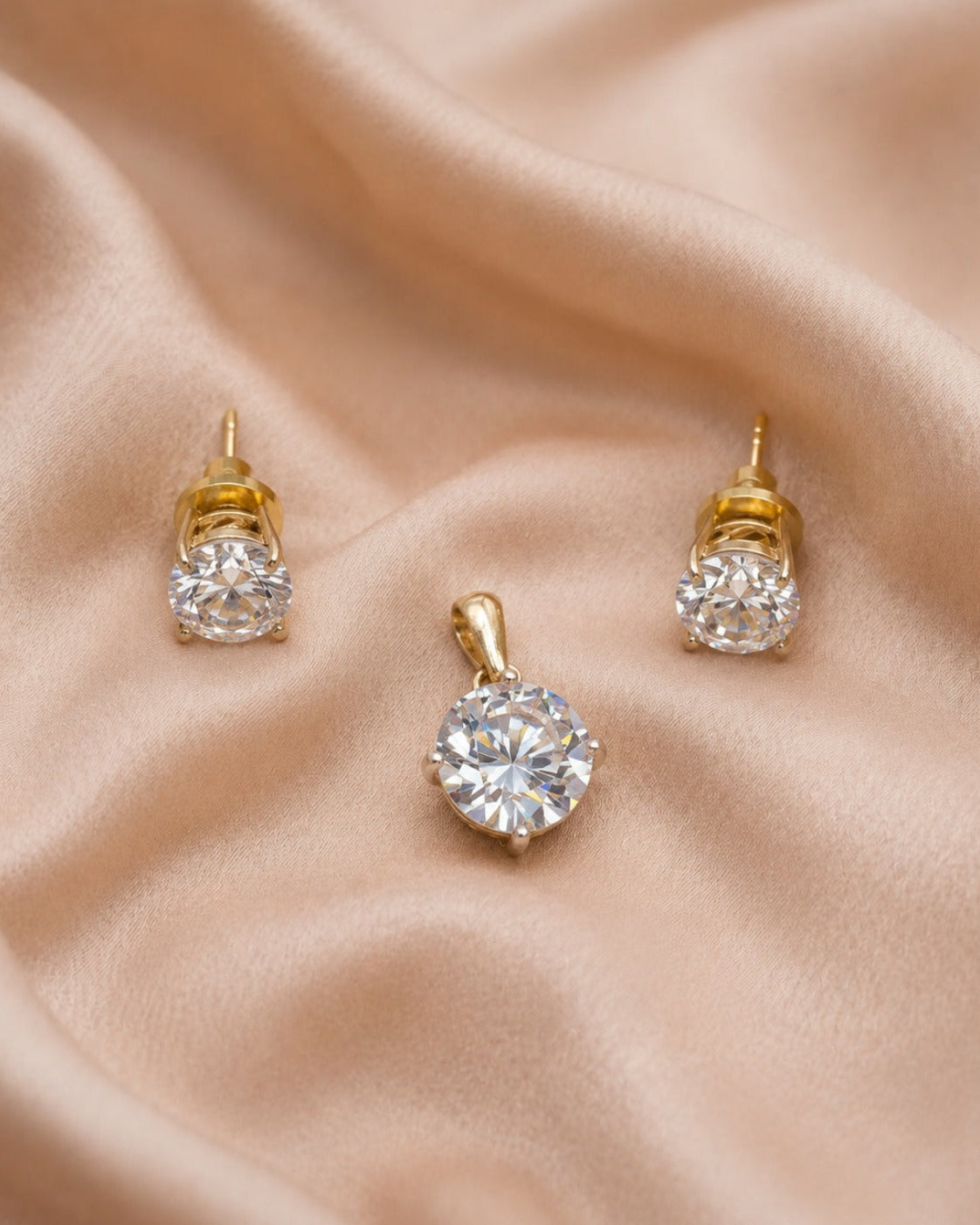 A pair of gold stud earrings and a matching pendant with clear round gemstones, arranged on draped beige satin fabric.
