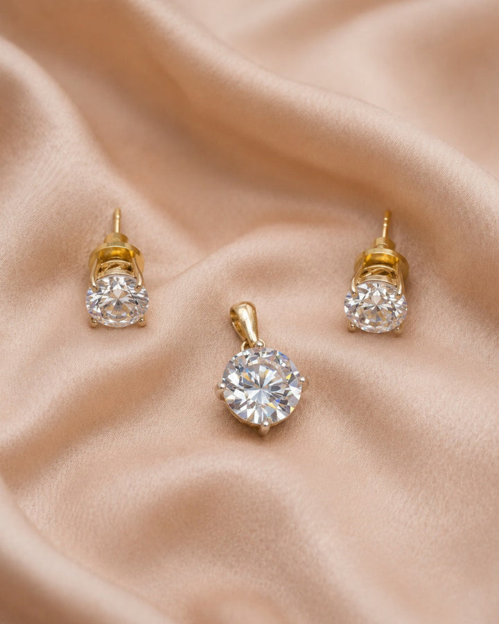A pair of gold stud earrings and a matching pendant with clear round gemstones, arranged on draped beige satin fabric.