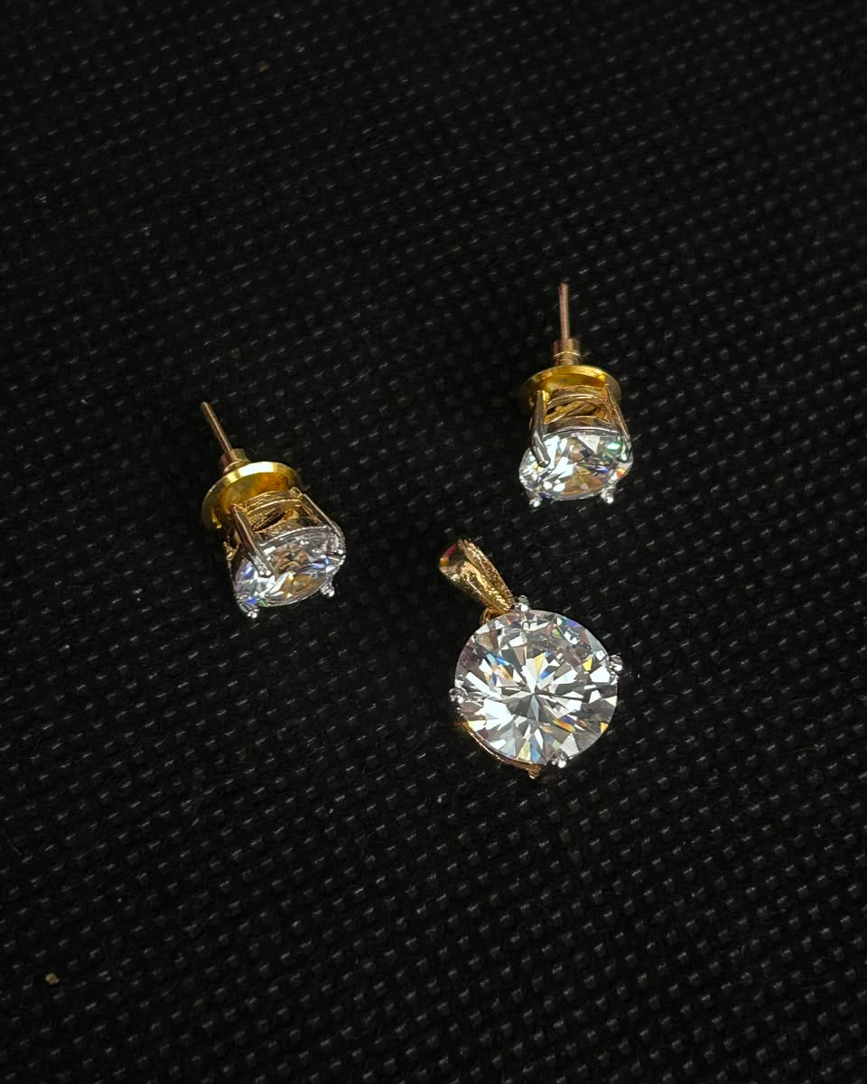 A pair of gold stud earrings and a matching pendant with large clear round-cut gemstones, displayed on a textured black background.
