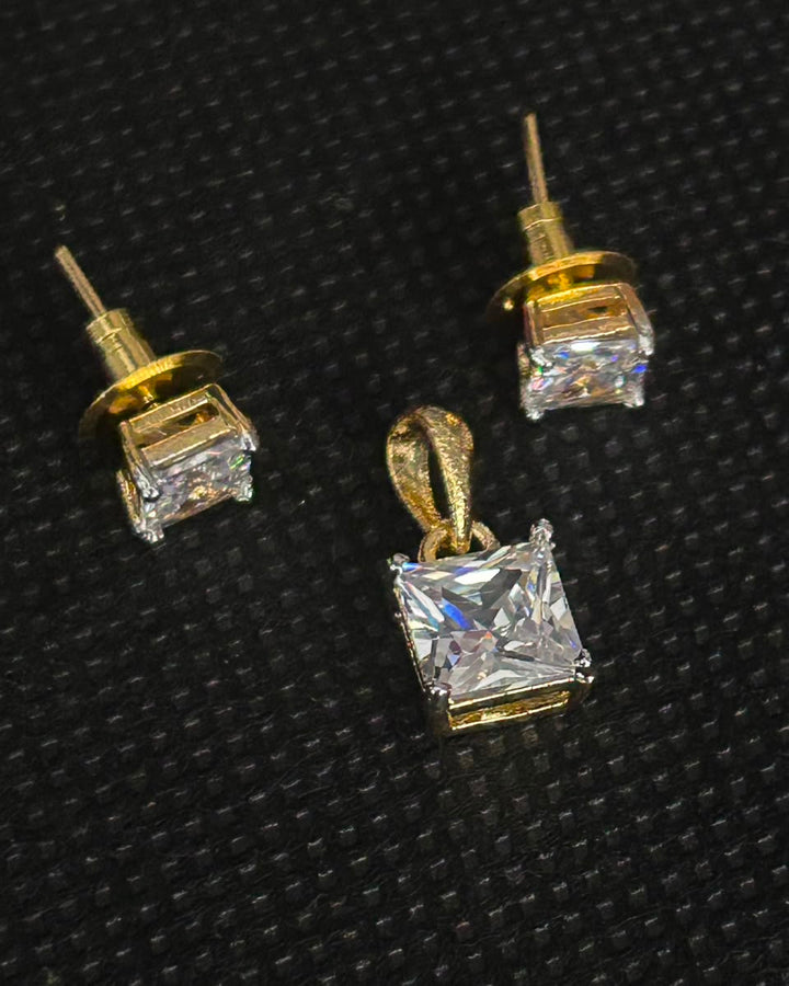 A pair of gold stud earrings and a matching pendant set with clear square princess-cut gemstones, displayed on a textured black background.