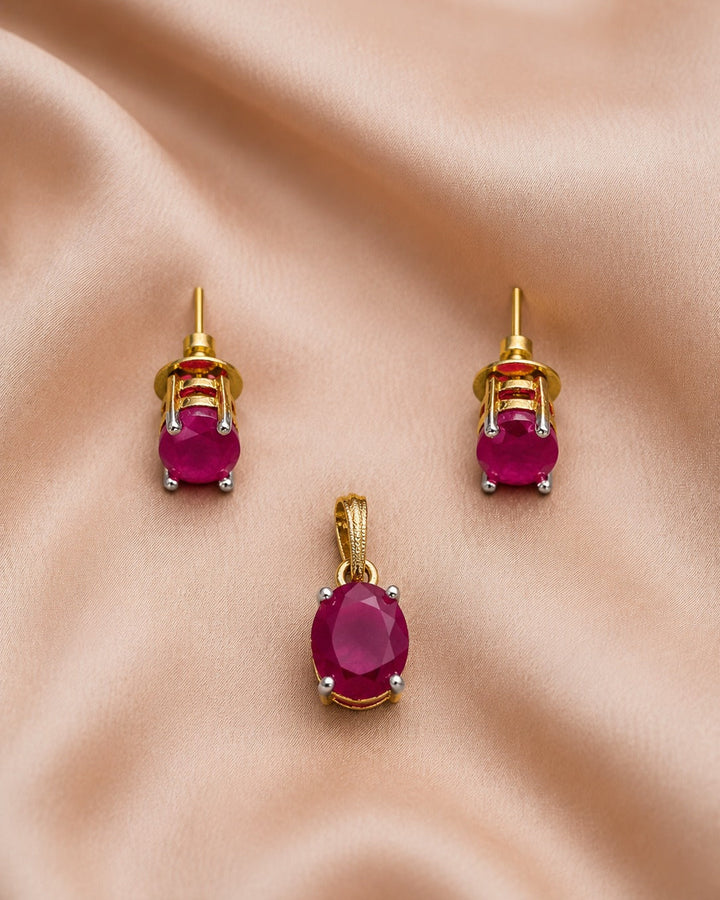 A pair of gold stud earrings and a matching pendant set with oval pinkish-red gemstones, arranged on draped satin fabric.