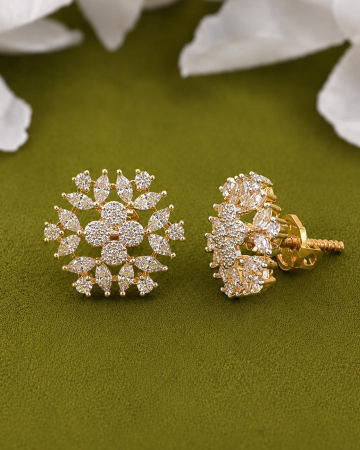 A pair of gold stud earrings decorated with clear pavé gemstones in a floral snowflake design, displayed on an olive green textured surface with blurred white flower petals in the background.