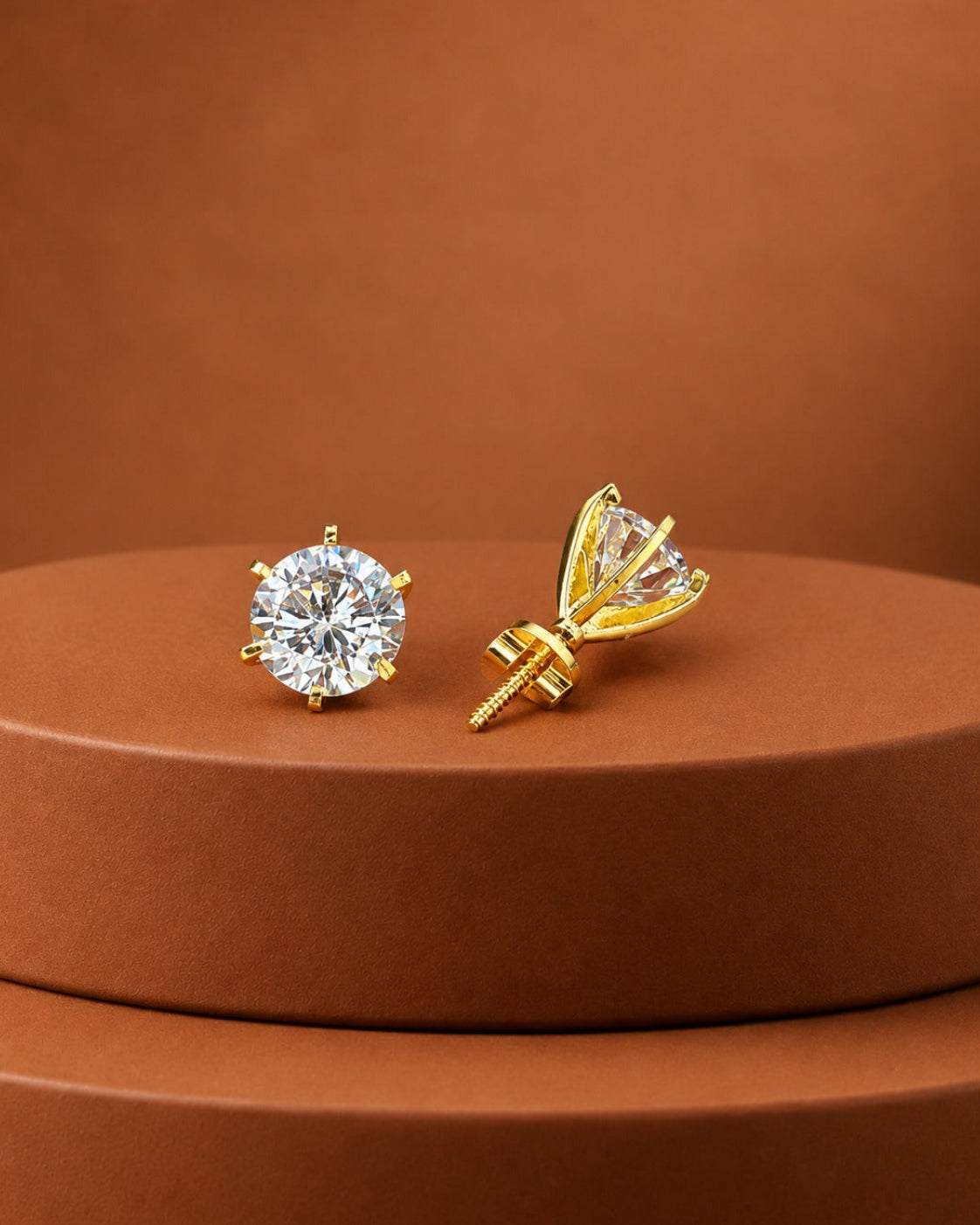 A pair of gold stud earrings displayed on a round brown pedestal. One earring is shown front-facing as a round brilliant-cut clear gemstone in a gold setting, and the other is shown from the side/back, revealing a prong design and screw-back post.