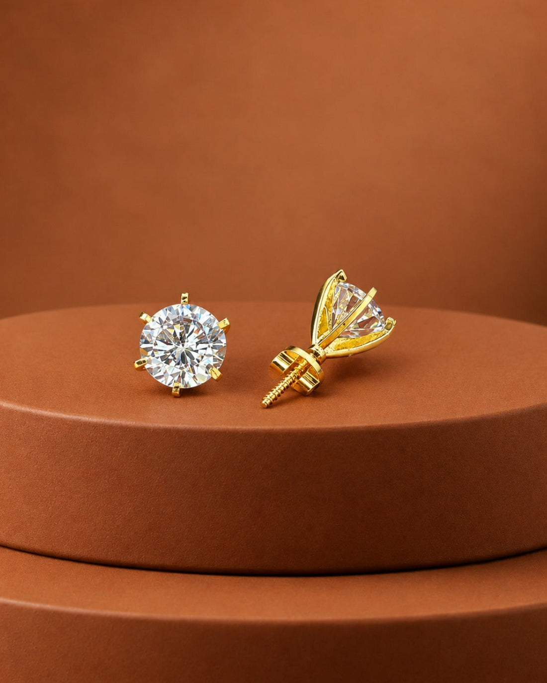 A pair of gold stud earrings displayed on a round brown pedestal. One earring is shown front-facing as a round brilliant-cut clear gemstone in a gold setting, and the other is shown from the side/back, revealing a prong design and screw-back post.